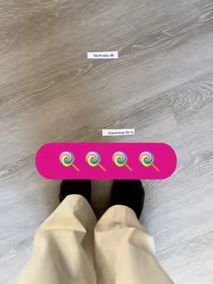 The image displays a top-down view of a person's legs and feet, standing on a light-colored wooden floor. The person is wearing beige pants and black socks. In the middle of the frame, there is a pink banner with four lollipop emojis. Above the banner are two white rectangular cards with text. The top card says 'MyProtein 48', and the bottom says 'Ginatricot 20%'. The lighting appears to be natural, and the overall composition is simple and straightforward.