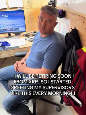 A man in a blue t-shirt is seated on a black office chair in front of a desk. A computer monitor is visible on the desk, and text in the image reads "I WILL BE RETIRING SOON FROM XRP...SO I STARTED GREETING MY SUPERVISORS LIKE THIS EVERY MORNING!!!"