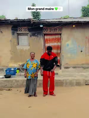 A man and an older woman stand in front of a humble building. The man, dressed in a red and black tracksuit, appears to be the focal point. The woman is wearing a blue and yellow patterned shirt, and a gray skirt. The background includes a weathered building with a corrugated metal roof, and simple, rural surroundings.