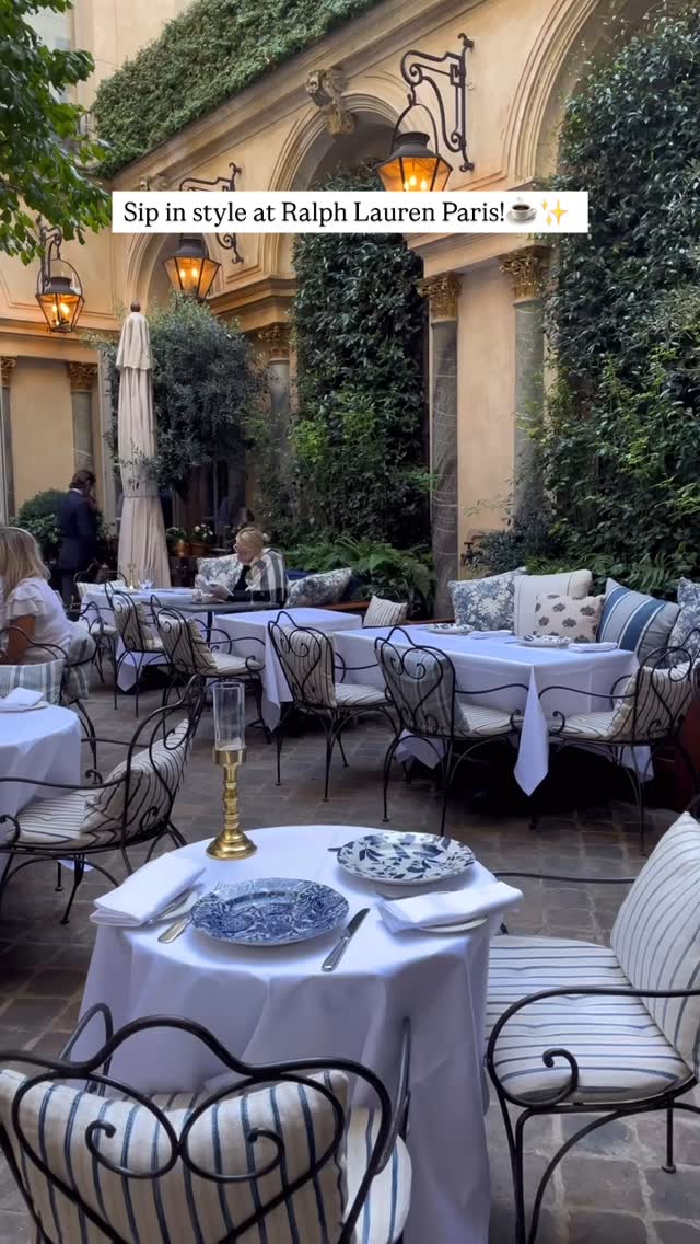 An outdoor shot of a cafe courtyard at daytime. The space is enclosed by stone walls with archways and lush greenery. Tables with white tablecloths are set with plates, cutlery, and blue and white patterned plates. Chairs with metal frames and striped cushions are arranged around the tables. Further in the background, people are seated at tables. The image has a natural light and a warm atmosphere. The text overlay says: Sip in style at Ralph Lauren Paris!