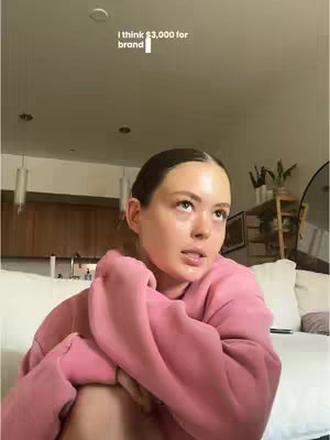 A woman with light skin looks upwards, seemingly deep in thought, while sitting on a white couch. She wears a light pink hoodie. The background includes a section of cabinets, some lit-up decor, and a few plants. A text overlay at the top says, "I think $3,000 for brand".