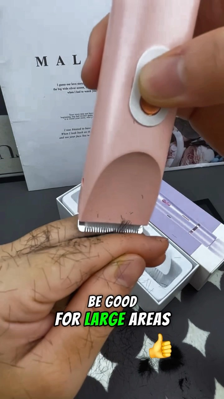 A close-up shot shows a person's hand, with the fingers holding a pink and white electric shaver. The shaver is shaving the fingertips of the hand, and small cut hairs are visible where the shaver blade has traveled. Behind the hand, parts of a white box and another box with a purple design are visible. In the background is a white paper with the word “MAL” on it.