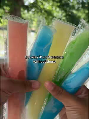 A person's hands holding a bundle of ice pops of various colors. The background is blurred, suggesting an outdoor setting.