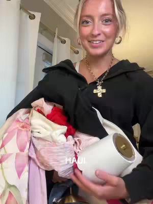 A young woman with blonde hair, wearing a black hoodie and a gold necklace, holds a pile of clothes and a white cylindrical object, likely a vacuum cleaner brush or a cleaning tool. The background reveals white curtains and a glimpse of a room.