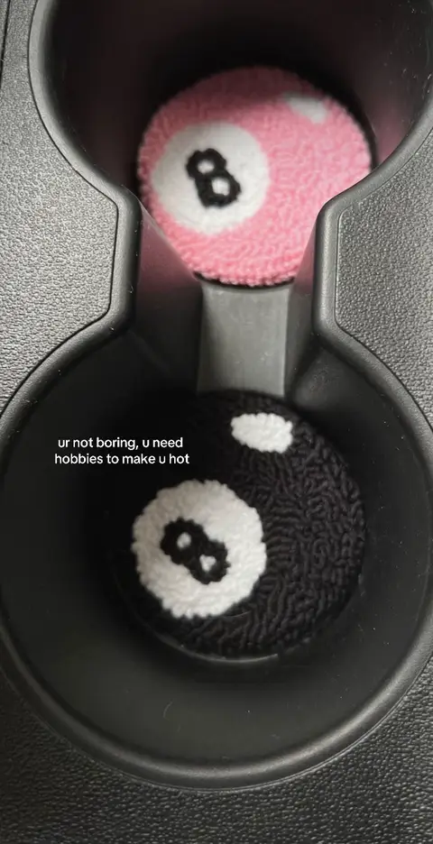 The image shows a car's cup holder with two 8-ball coasters. The top coaster is pink, the bottom one is black. Text overlayed reads 'ur not boring, u need hobbies to make u hot'.