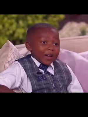 A young Black boy wearing a formal outfit (vest, tie, shirt) and looking up with a surprised or questioning expression while seated on a couch.