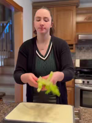 A woman stands in a kitchen, holding a bunch of celery. She is wearing a dark cardigan over a green top. The lighting is natural, and the composition is a medium shot, focusing on her upper body and the kitchen environment.