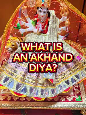 The image shows a close-up of a Hindu deity statue, likely a goddess, adorned with elaborate clothing and jewelry. The statue is positioned with a devotional offering in front of it. In the foreground, there is a hook text placed on top of the statue.