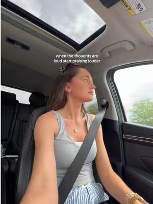 A woman is seated in the driver's seat, looking out the window to her right, with the sun roof open overhead. The image is taken from a low angle, giving a sense of being in the car with her. The car's interior is visible, including the black seat and a portion of the dashboard. The sky is overcast, but the car interior is brightly lit. A hook text, in white, is at the top left of the windshield.