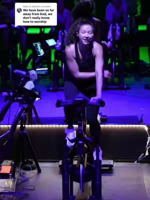 A woman is shown riding a stationary bike in a spin studio. She is in the center of the frame, wearing a black tank top and leggings. The background is blurred, showing other people on bikes and equipment. The lighting is dim, with blue and green hues.

The image is a medium shot, focusing on the woman's upper body and face. She is smiling and appears to be enjoying the workout. There is a microphone in front of her face.
