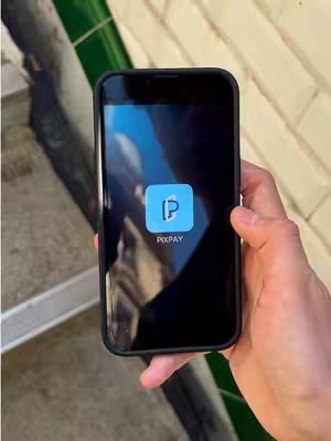 A hand holding a smartphone that displays the Pixpay app, set against a background that includes outdoor features like stone. The phone's screen is the focus, with the app logo prominent. The overall composition has clean lines and a modern feel.