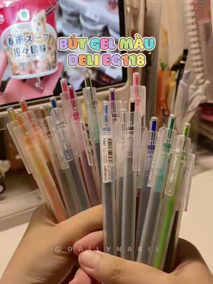 A person's hands are holding a bundle of colorful gel pens, with a blurred background that includes a screen showing a food product. The hands, which appear to be of a person with red nail polish, are in the foreground, with the pen tips clustered together. The pens are different colors, with some clear and some translucent. In the background is a person holding a take-out food cup and a blurred computer screen.