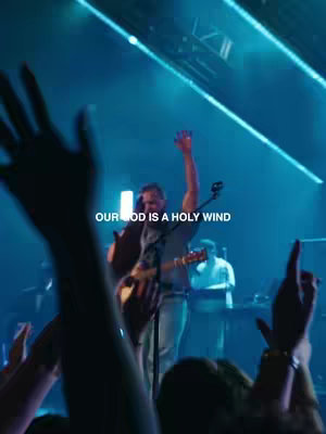 A live music performance on stage with a band. A man with a guitar is singing with his arm raised, and many hands of the audience are up as well.