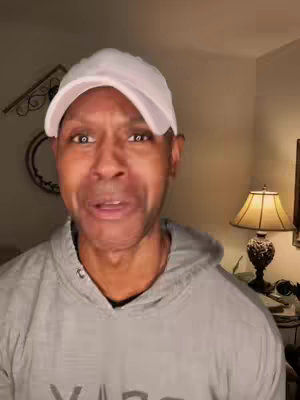 A medium shot shows a Black man looking directly at the camera with a slight wince. He wears a white baseball cap and a gray hoodie, the hood down. The background includes a lamp with a lampshade casting a warm light, a decorative wall hanging, and parts of a wall and a possible framed mirror. The lighting is soft.