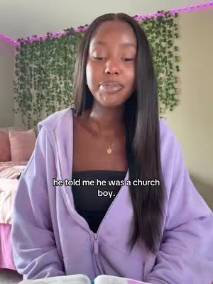 A young Black woman with long, dark hair is centered in the frame. She wears a lavender zip-up hoodie and a black strapless top, with a gold necklace. Behind her, a bedroom wall is decorated with hanging greenery. A text overlay reads, 'he told me he was a church boy.' The overall composition is well-lit and informal, suggesting a casual video.