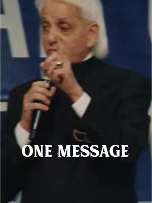 A man with white hair and a black suit is holding a microphone and speaking to the audience. The man is wearing a gold ridged ring on his right ring finger. The background is out of focus.