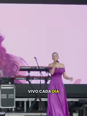 A woman in a purple dress is singing on a stage. Behind her is a large pink backdrop, and a keyboard is visible in front of her