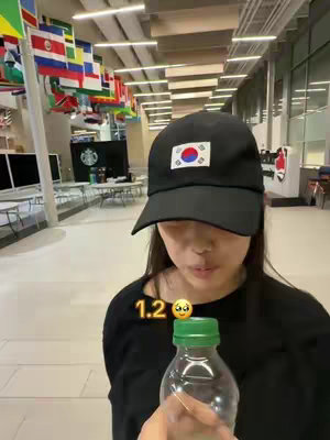 A young woman wearing a black cap with a flag on it is holding a water bottle looking at the camera with a concerned expression. The background includes a hallway with flags and a Starbucks sign.