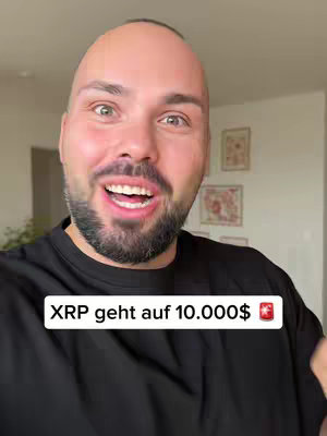A close-up selfie of a fair-skinned man. He has a short, trimmed beard, and is smiling with his mouth open, showing his teeth. The background is a light-colored wall with framed pictures. A text overlay in a black rectangle reads 'XRP geht auf 10.000$' with a flag next to the text.