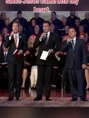 The image shows three men on a stage, each holding a microphone. They are dressed in suits and appear to be singing or speaking in front of a background of other people. The setting looks like a church or auditorium, with a choir visible in the background.