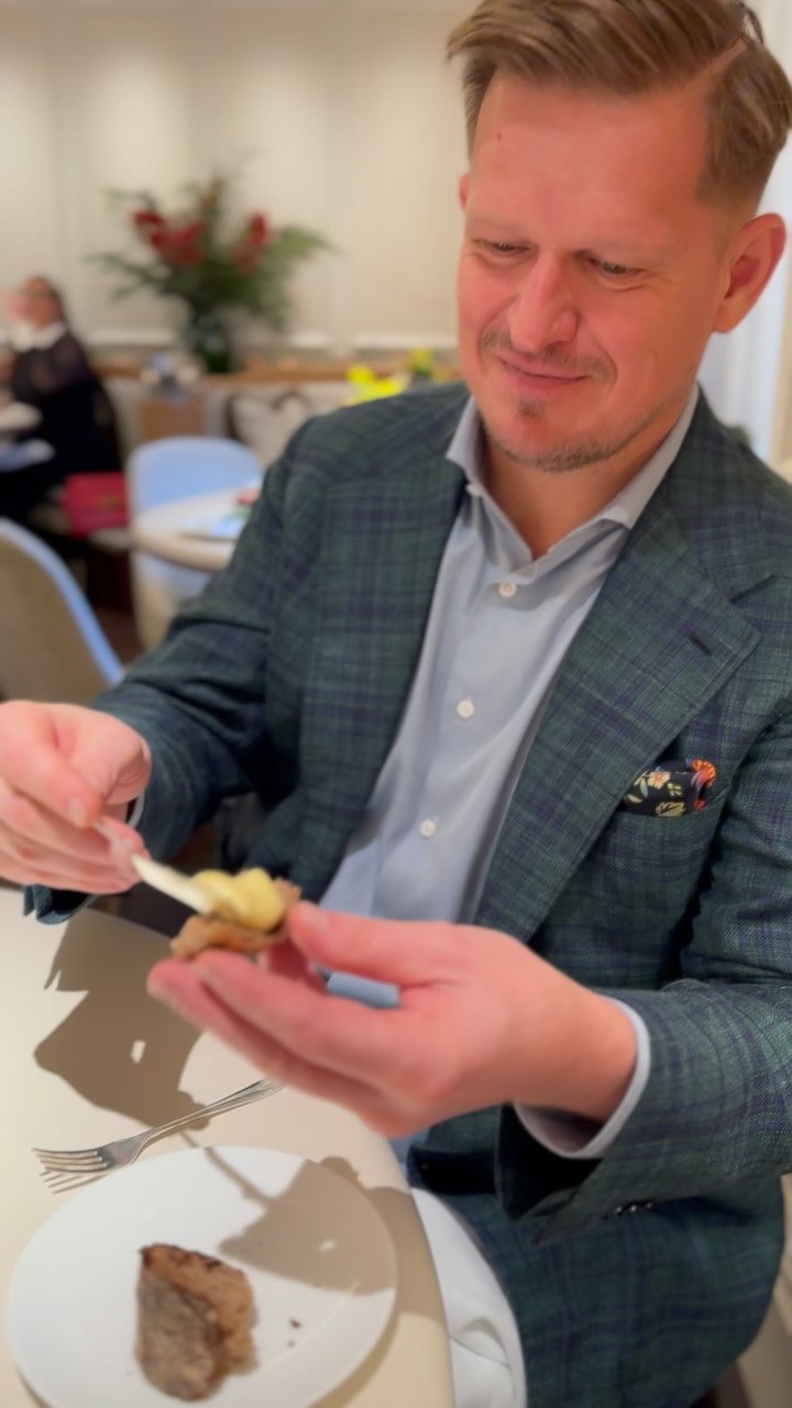 A man in a green and blue plaid blazer is holding a small piece of food, possibly a hors d'oeuvre, with a small wooden utensil. He is seated at a white table in a restaurant, with a plate of food to his side. The background is blurred, suggesting the setting is a fine dining establishment.