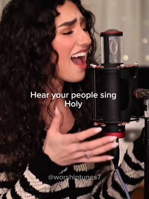 A young woman with dark curly hair sings into a microphone. The image is a close-up, highlighting her face with visible emotion. The woman is wearing a black and white striped top, and a silver ring is visible on her finger. The background is blurred, suggesting a performance setting or studio. Text overlayed on the image reads, 'Hear your people sing Holy'.