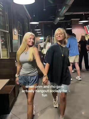 Two young women, holding hands and walking towards the camera, are shown in a dimly lit indoor space. Both women are wearing casual attire and have long blonde hair. The image is framed from a slightly low angle, with muted lighting and visual elements suggesting a casual, social setting.