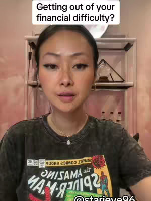 A woman with fair skin and dark hair is centered in the frame. Above her head, in a white box with a black border, is the text 'Getting out of your financial difficulty?'. She is looking directly at the camera. Her dark hair is shoulder-length. She has subtle makeup on her face. She’s wearing a black t-shirt featuring a comic book character, and a gold necklace with a small pendant. Behind her is a shelf filled with trinkets, and the walls are a soft pink. The lighting is bright and even.