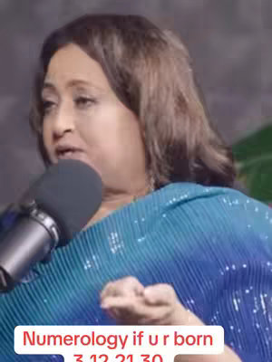A woman with dark hair wearing a blue and teal top is holding a microphone and speaking. A text overlay in a red box sits below her.