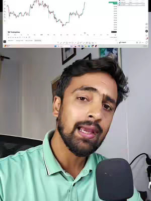 A man with a beard, wearing a green shirt, looks towards the camera. Behind him, a computer screen displays a stock chart.
