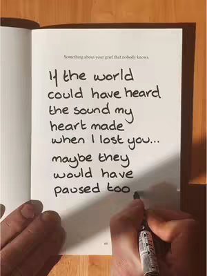 A hand holding a black pen is writing text on a white page in an open book. The page contains handwritten text in black ink with the title 'Something about your grief that nobody knows.' The text reads 'If the world could have heard the sound my heart made when I lost you... maybe they would have paused too.' The background visible is a wooden surface.
