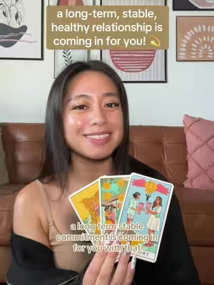 A woman smiles and holds three tarot cards, with text overlays above and below. The background includes a brown couch and artwork.