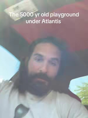The cover image presents a close-up of a man in the front seat of a vehicle, likely a car, with the camera angled from below. The man has long, dark hair and a full beard, suggesting a mature appearance. He is wearing a white shirt that contrasts with the darker interior of the car. Above him, a text overlay reads, 'The 5000 yr old playground under Atlantis,' set against an obscured background.