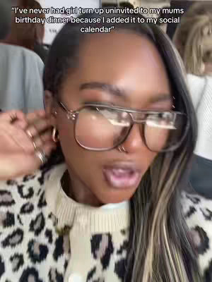A close-up shot of a woman with dark skin wearing large, square-framed glasses, and a leopard print sweater. Her mouth is slightly open, and her expression suggests surprise or amusement. The background is blurred, with hints of other people and objects.