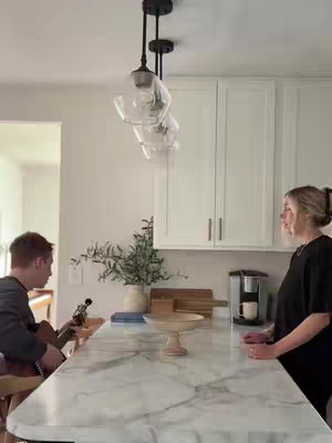 The cover image captures a light-filled kitchen scene. Two individuals are present—a man on the left playing a guitar while seated, and a woman on the right, standing and facing toward the man. The setting includes a marble-topped island, white cabinets, and hanging lights. The background is slightly blurred, emphasizing the subjects and the warm, inviting kitchen aesthetic. The color scheme is neutral, with the focus on the individuals and their interaction.