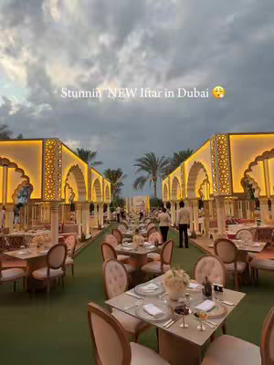 An outdoor setting at dusk with tables set for a meal. There are decorated archways and palm trees. The sky is filled with clouds.