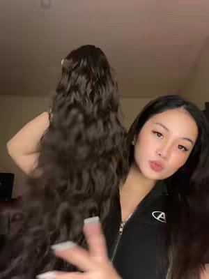 A young woman with long, dark hair stands in front of a dark background. She is holding a section of wavy, dark hair in front of her. She is looking at the camera with a slight pout and expression. The lighting is soft, and her face is illuminated.