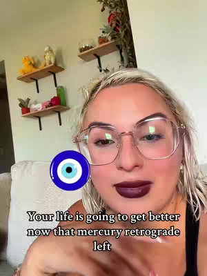 A selfie-style video cover features a woman with blonde hair, wearing large-framed glasses, and dark lipstick. An eye-shaped charm floats near her face. Shelves with decorations are behind her. Text overlays are present at the bottom of the frame.