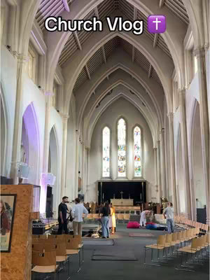 The image is a wide shot of the interior of a church. The architecture is classic, with high vaulted ceilings and tall, arched windows. Stained glass windows are visible in the background. The floor is covered with a black covering. There are several people in the church, some seemingly setting up and others standing or sitting. The image overall is bright and airy