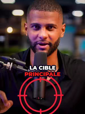 A man is centered in the frame, holding a microphone with both hands. In the foreground, there's text reading 'LA CIBLE PRINCIPALE', set against a red target symbol. The background is slightly blurred, suggesting a studio or indoor setting.
