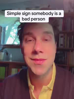 A man with short, dark hair is centered in the frame, slightly smiling. He is wearing a yellow shirt under a dark navy or black cardigan. The background features a blurred indoor setting, including bookshelves and a window. A white bar at the top of the screen contains text.