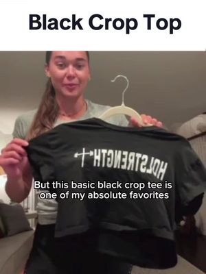 A woman is shown holding up a black crop top, revealing the shirt's design and positioning it in front of herself. She is in a well-lit indoor setting with text overlayed at the top and bottom of the screen..
