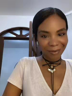A woman with dark hair and a fair complexion is centered in a medium shot. She wears a white top and a black choker with a heart pendant. The background consists of white walls and a wooden door. She is smiling, and the lighting appears to be natural.
