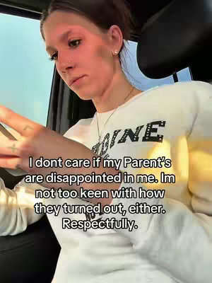 A woman is seated inside a car, angled slightly towards the camera. She's wearing a white sweatshirt with the word 'INE' visible on the front. The lighting is somewhat diffused. The car’s interior creates a simple backdrop. The woman has light skin, dark hair pulled back, and appears to be in her mid-20s to early 30s. The background is slightly blurred, placing focus on her.
