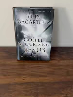 An image of a hardcover book titled "The Gospel According to Jesus" by John MacArthur, on a wooden surface. The book is shown upright, with the title prominent against a background of storm clouds. The wooden surface seems to be a table or desk.