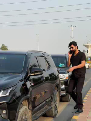 A man in a black polo shirt and black pants stands outside, talking on a phone. He is partially framed by two black SUVs parked nearby. The background shows a clear sky, road, and some greenery.