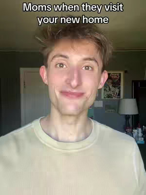A young man with short brown hair, wearing a light yellow shirt, smiles directly at the camera. Above him is white text that reads, 'Moms when they visit your new home.' The background includes a glimpse of a framed picture on a wall and a lamp.