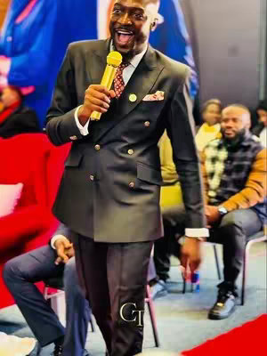 The image showcases a man giving a speech on a stage. He is dressed in a dark double-breasted suit and is holding a gold microphone. Behind him, there are other seated people and what seems like a red couch. The lighting is bright and the man appears to be energized.
