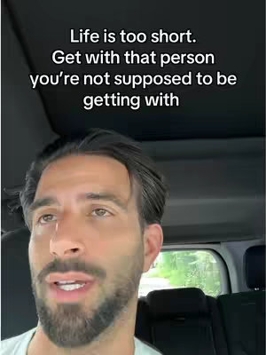 A close-up shot taken inside a car, focusing on a man with dark hair and a beard, who appears to be speaking. Above him, text overlays in a white font.