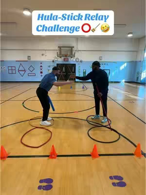 Two men are in what appears to be a gymnasium, participating in a relay race. They are facing each other with their arms outstretched in a passing motion. On the ground, there are orange cones, hula hoops, and markers to indicate the track.